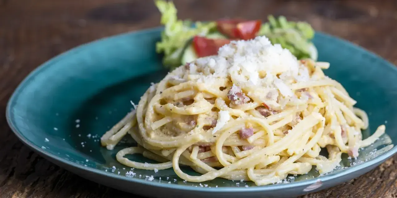 Porcja spaghetti carbonara posypana startym serem, podana na turkusowym talerzu z sałatką.