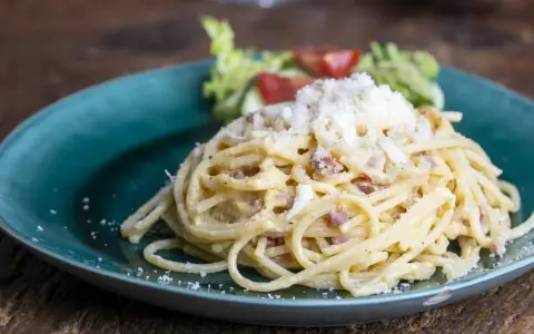 Porcja spaghetti carbonara posypana startym serem, podana na turkusowym talerzu z sałatką.