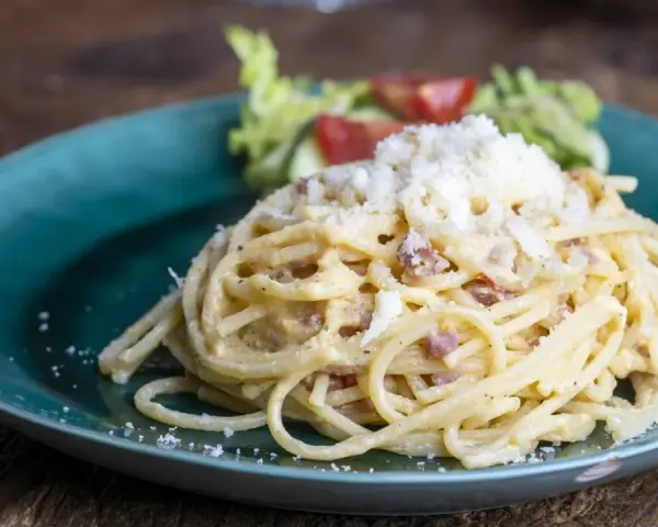 Porcja spaghetti carbonara posypana startym serem, podana na turkusowym talerzu z sałatką.