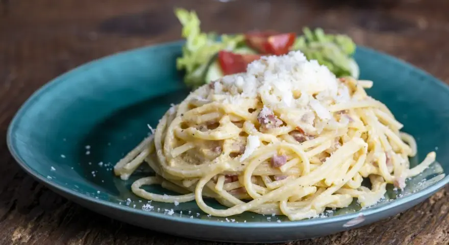 Porcja spaghetti carbonara posypana startym serem, podana na turkusowym talerzu z sałatką.